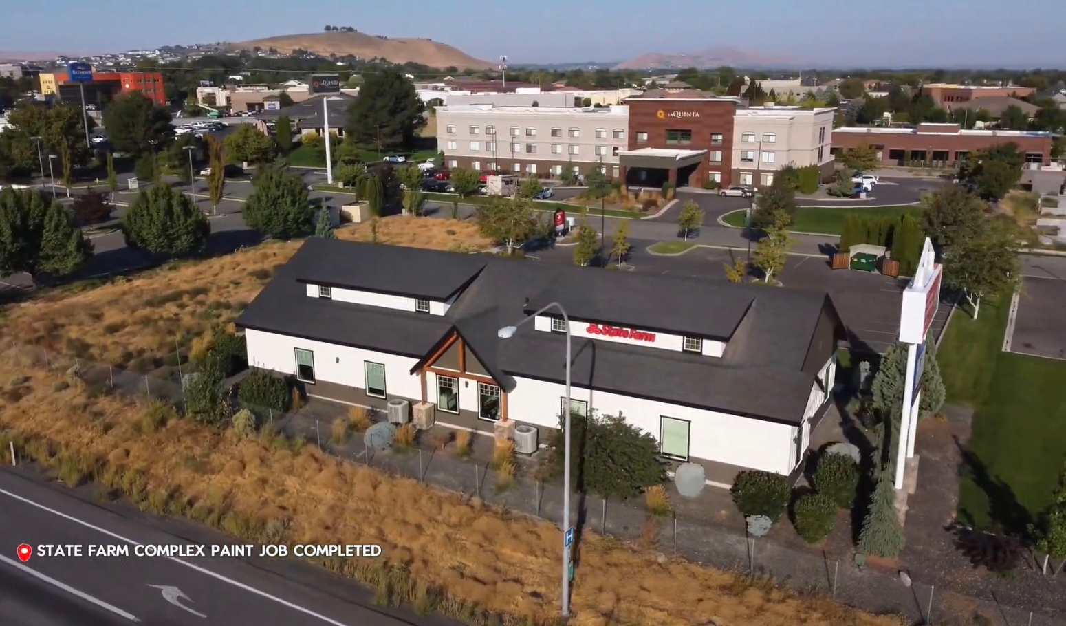 Aerial view of a commercial building with a dark roof and a hotel in the background, a sunny day.