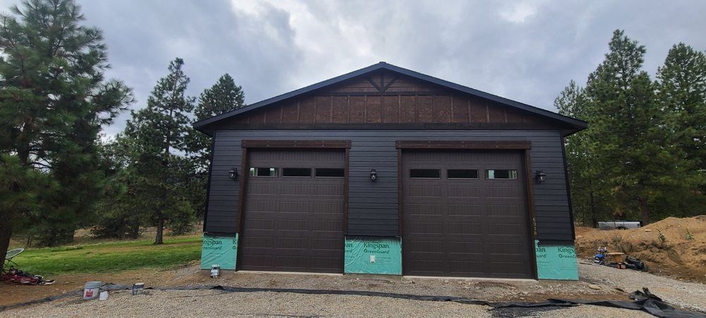 Two-car garage with brown doors and dark siding under a cloudy sky, surrounded by trees.