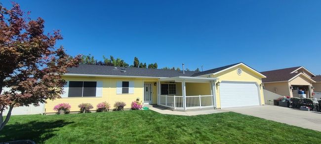 Yellow house with a garage and porch, surrounded by green grass and pink flowers under a blue sky.