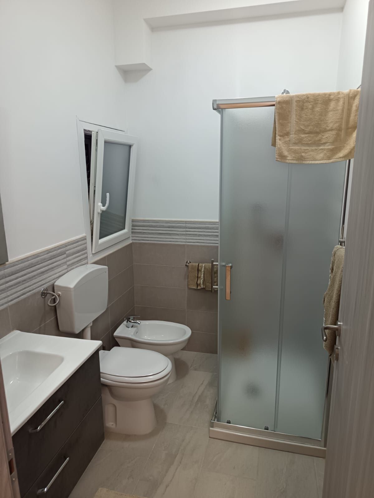 Piccolo bagno con WC, lavandino, box doccia satinato e specchio sopra piastrelle beige.