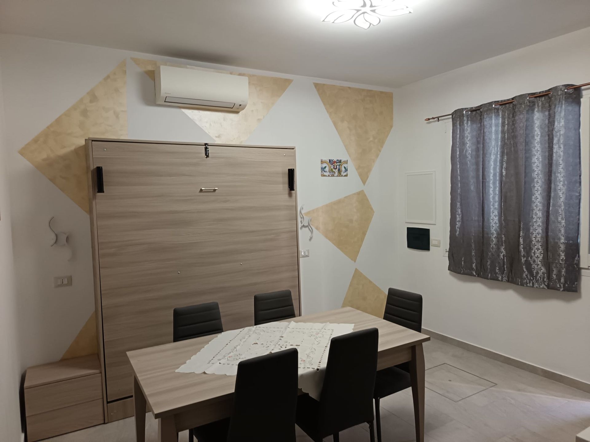 Piccola sala da pranzo con tavolo in legno, sedie nere, condizionatore a parete, tende e decorazioni murali beige.