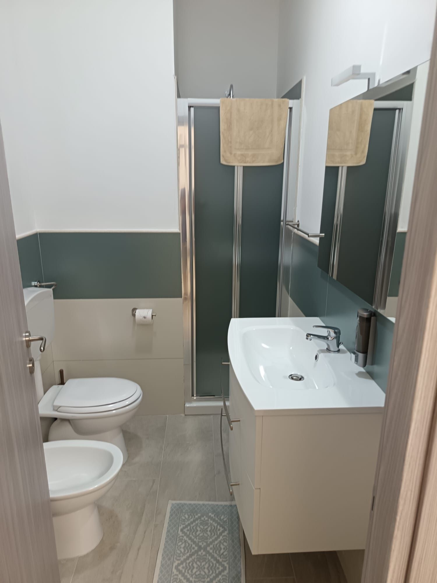 Piccolo bagno con WC, lavabo, doccia, specchio e portasciugamani nei toni del beige e del grigio.
