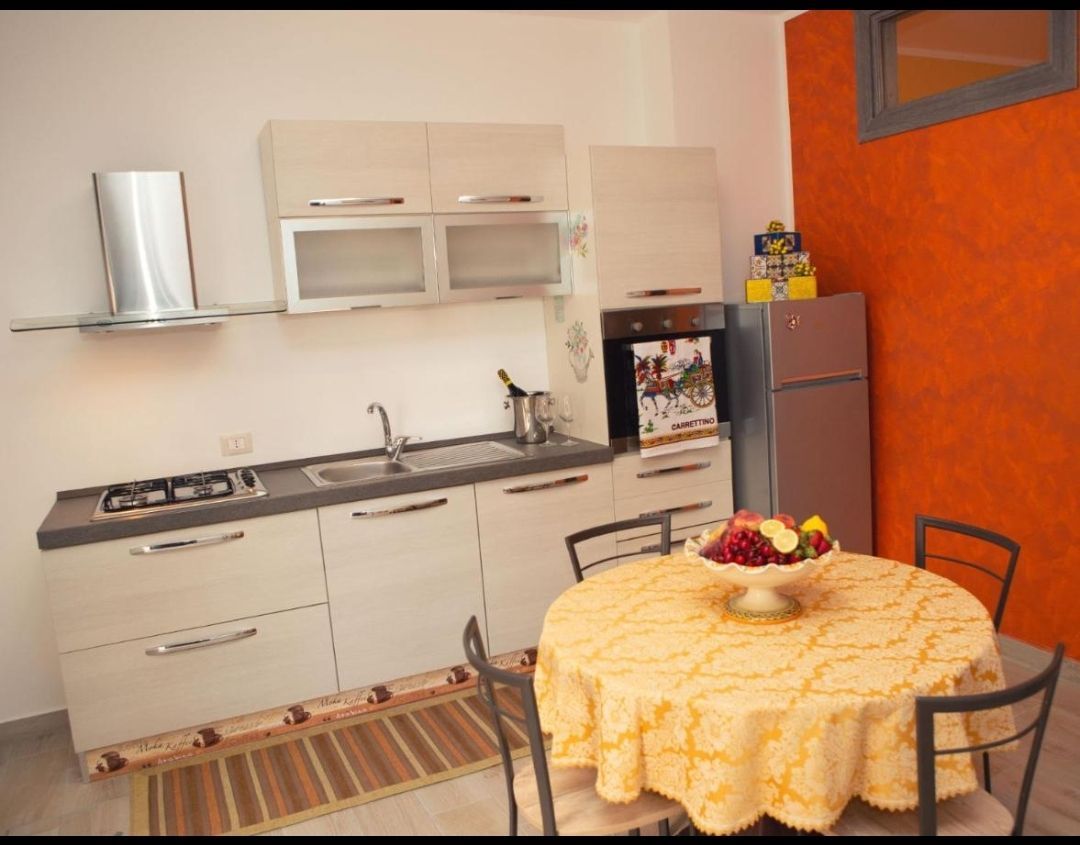 Cucina moderna con mobili bianchi, parete arancione e tavolo da pranzo rotondo con tovaglia gialla e fruttiera.