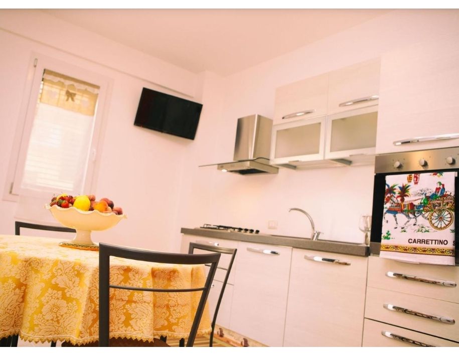 Cucina moderna e luminosa con mobili bianchi, piano cottura in acciaio inox, TV a parete e tavolo con tovaglia gialla.
