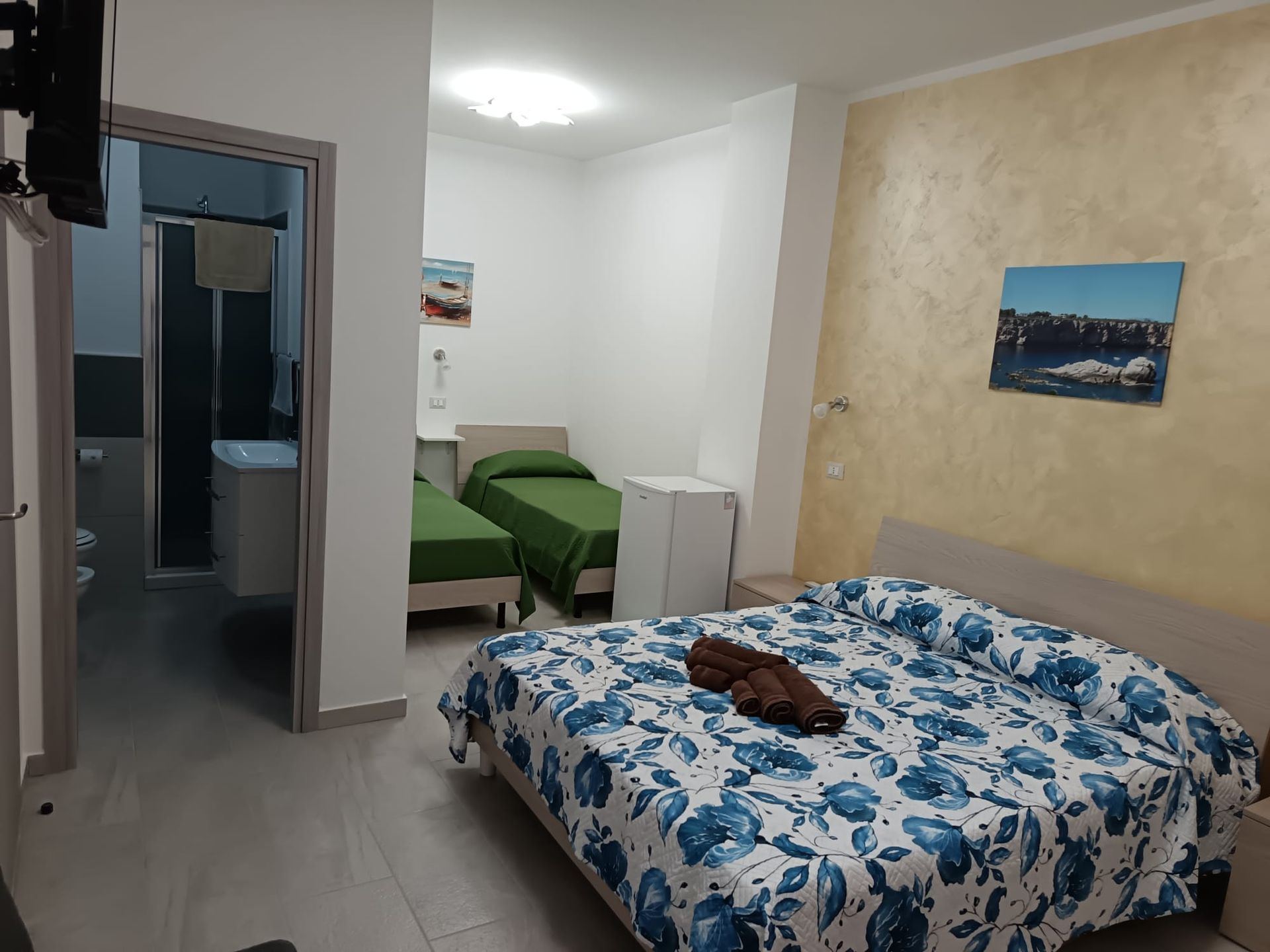 Piccola camera d'albergo con letto, sedia verde e porta aperta; lenzuola con motivi floreali blu e un quadro alle pareti.