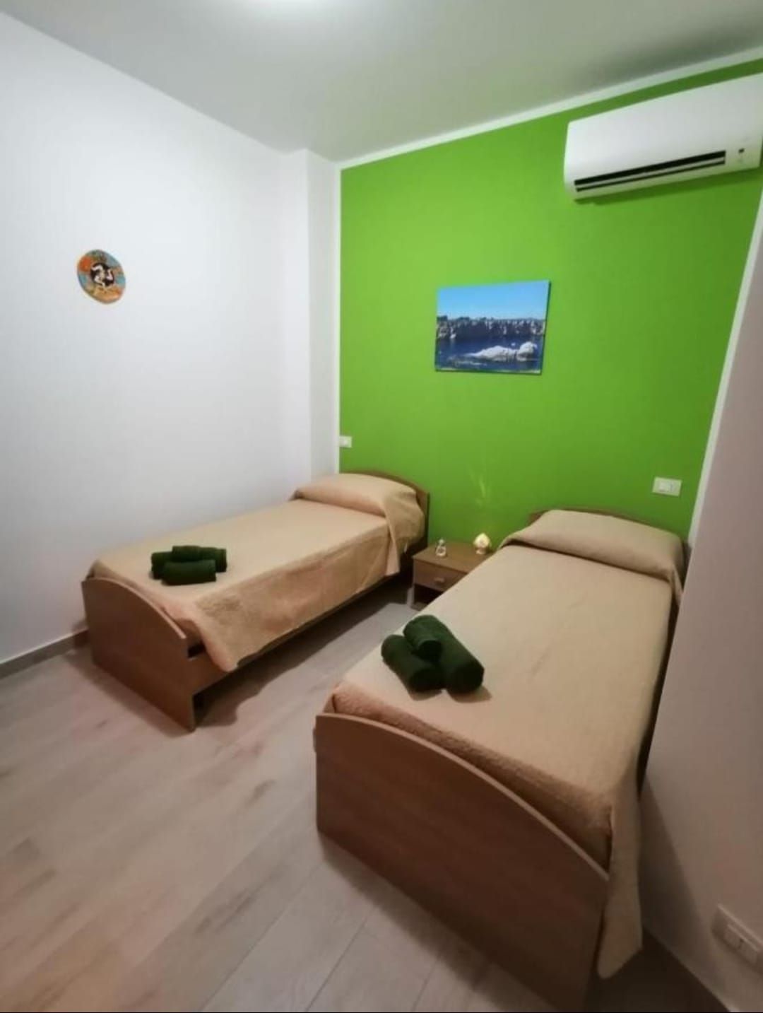 Piccola camera da letto con due letti singoli, una parete con dettagli verde brillante, quadri e un condizionatore d'aria.