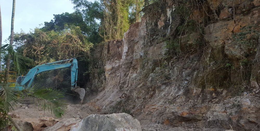 A Blue Excavator is Digging a Hole in a Rocky Area — Diggamen Civil Contracting in Tivendale, NT