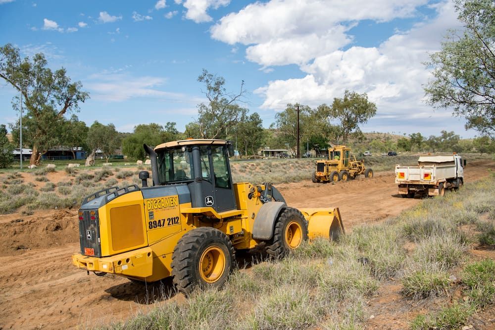 A Yellow Tractor Is Driving Down a Dirt Road Next to A Truck — Diggamen Civil Contracting in Jabiru, NT