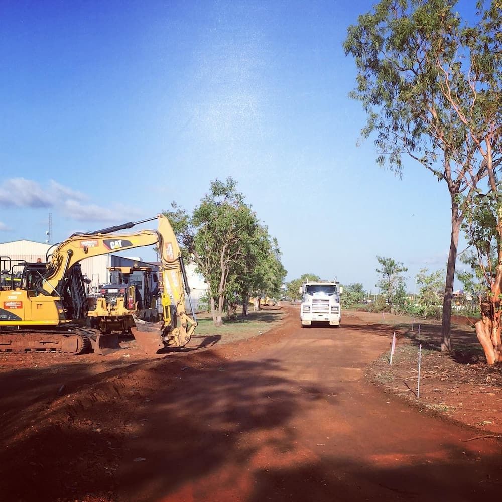 A Truck Is Driving Down a Dirt Road Next to A Yellow Excavator — Diggamen Civil Contracting in Tivendale, NT