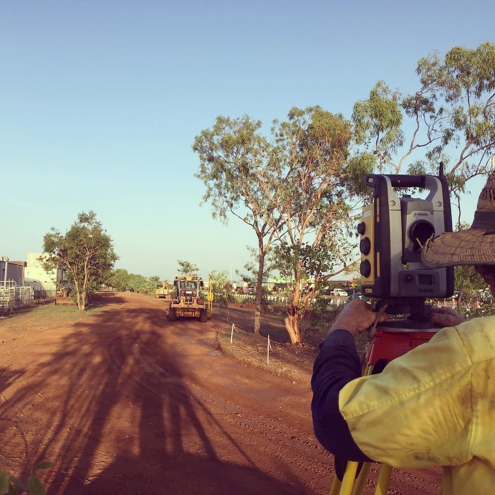 A Man in A Yellow Shirt Is Using a Total Station on A Dirt Road — Diggamen Civil Contracting in Palmerston, NT