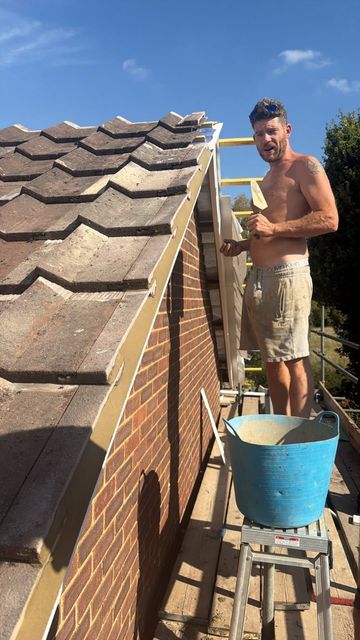 Worker on a roof cleaning or repairing shingles beside a brick wall and blue bucket.
