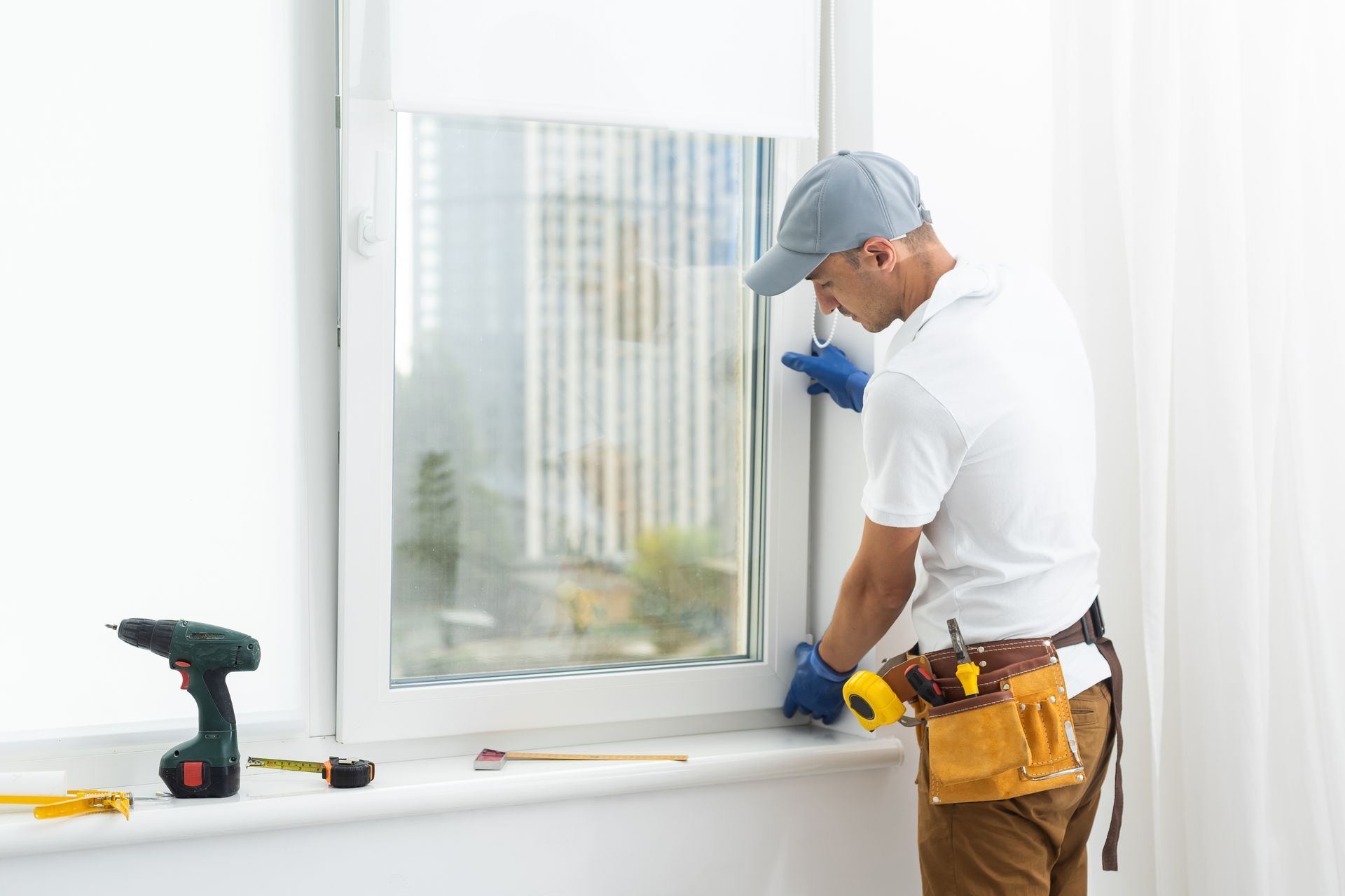 Man in work clothes installing window; tools on sill.