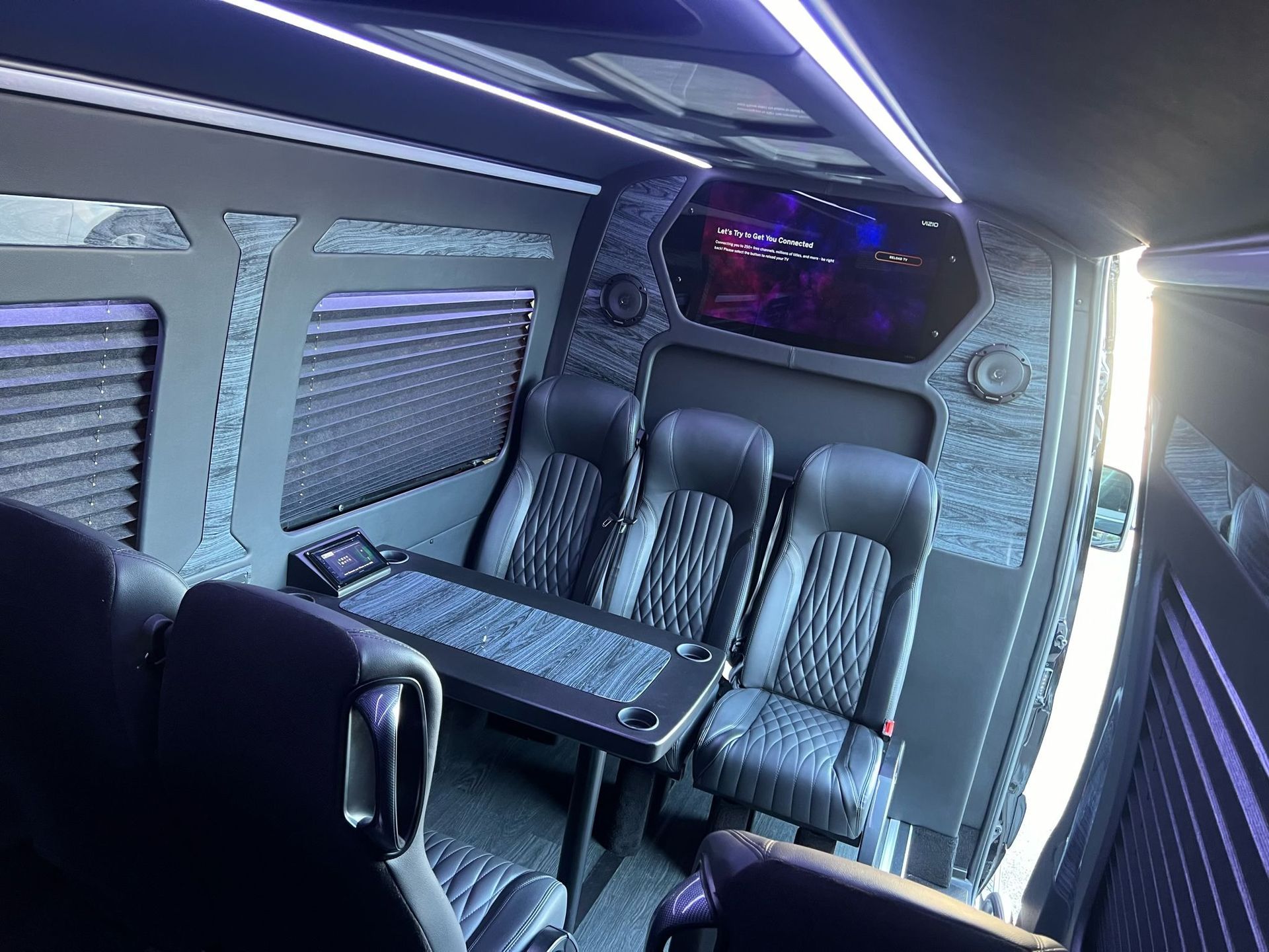 Interior of a luxury van with black leather seats, a table, and gray paneling with blue accent lighting.