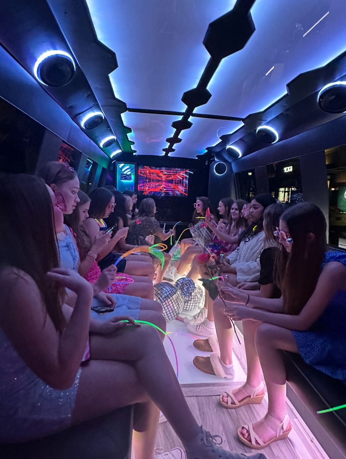 Teenagers in a limousine with neon lights; celebrating, sitting on benches, some on phones.