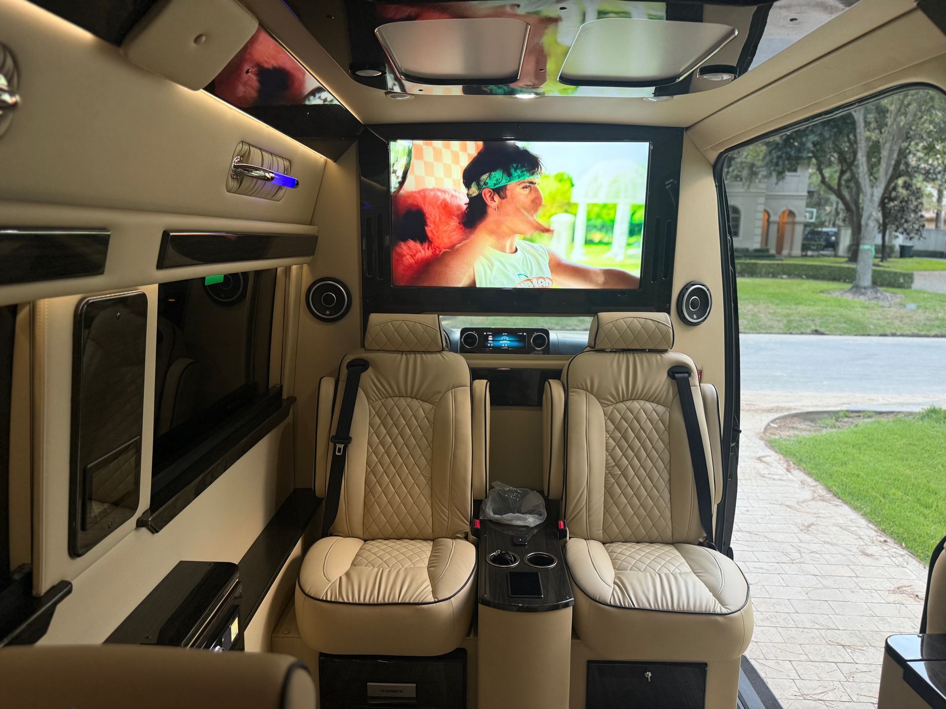 Interior of a luxury van with two beige leather seats, a large TV, and ambient lighting.