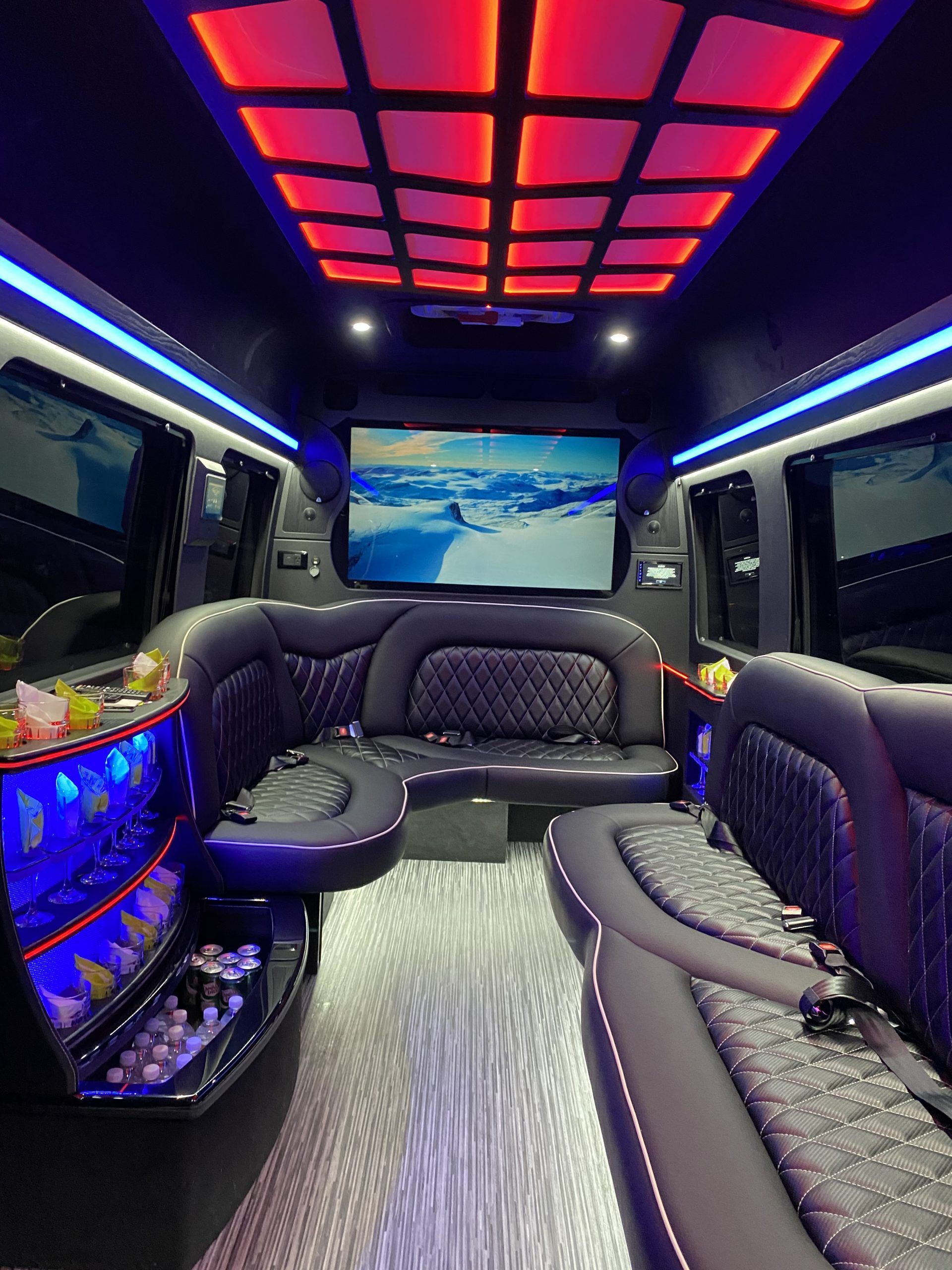 Interior of a luxury black limousine with blue and red ambient lighting, large screen, and plush seating.