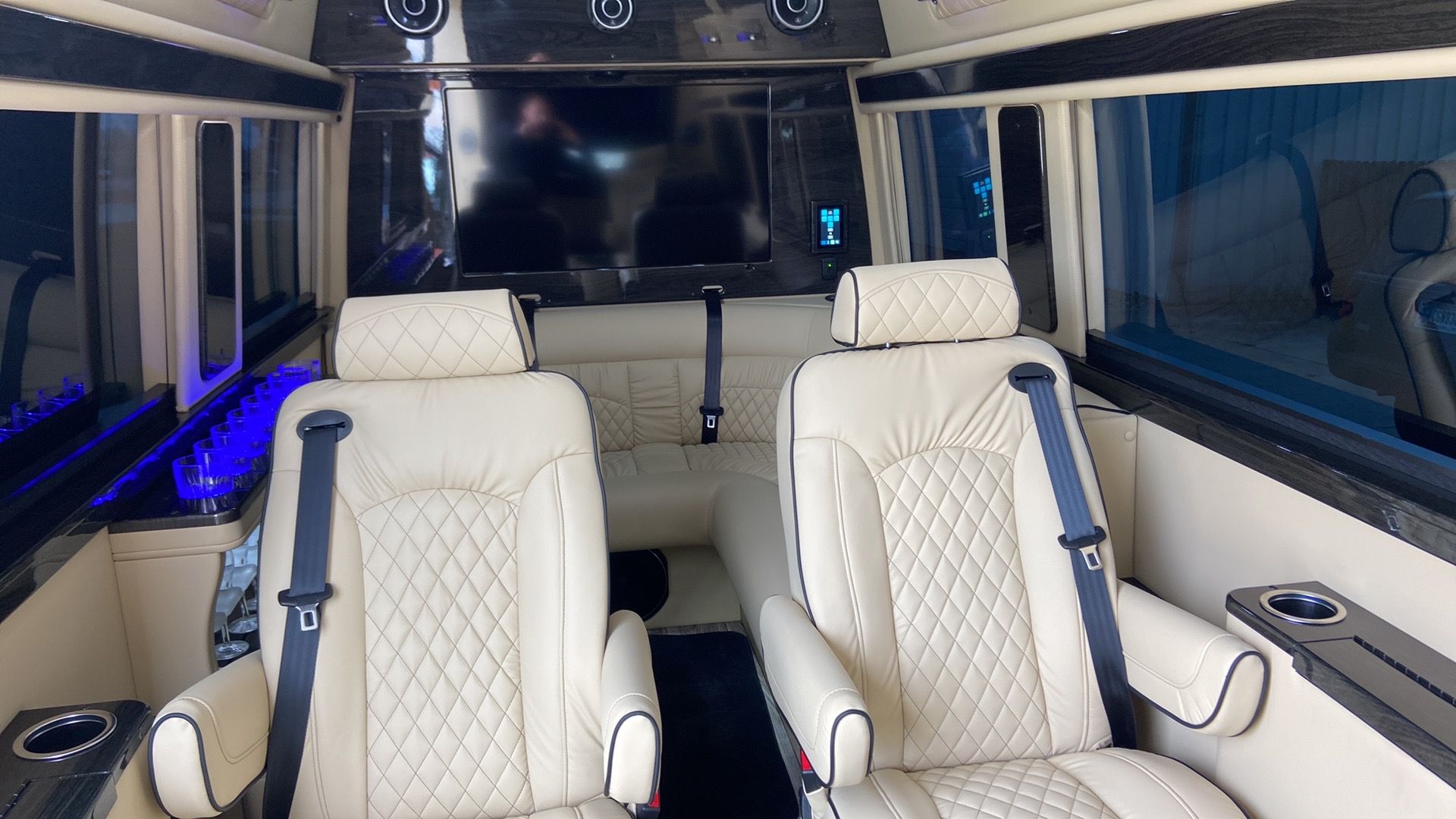 Interior of a luxury van with cream-colored quilted leather seats, a TV, and blue ambient lighting.