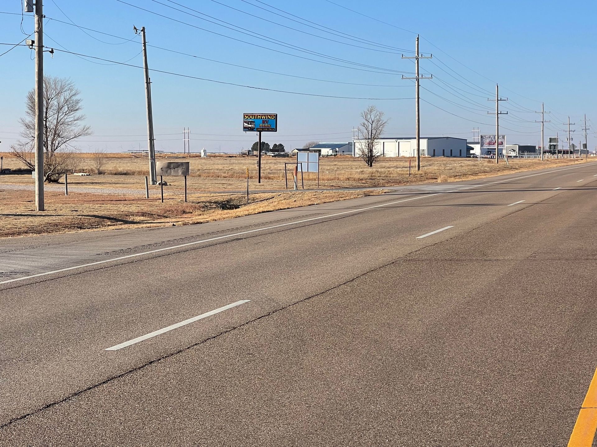 Empty rural road beside dry fields and utility poles under a clear blue sky.