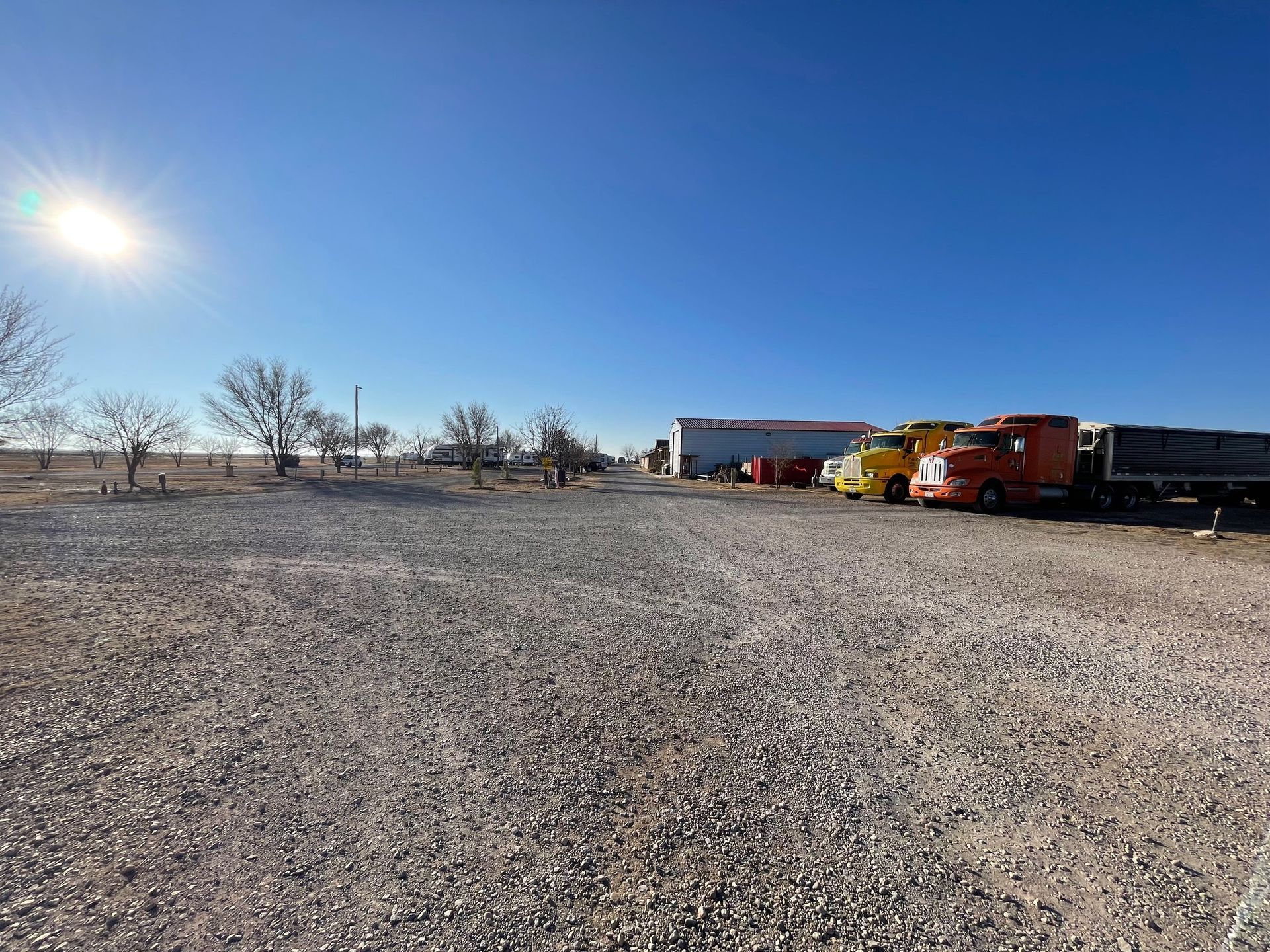 Wide gravel lot with a red-orange truck and small buildings under a bright blue sky