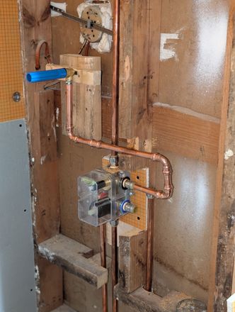 Exposed wall plumbing with copper pipes, valves, and a blue handle near a shower valve box