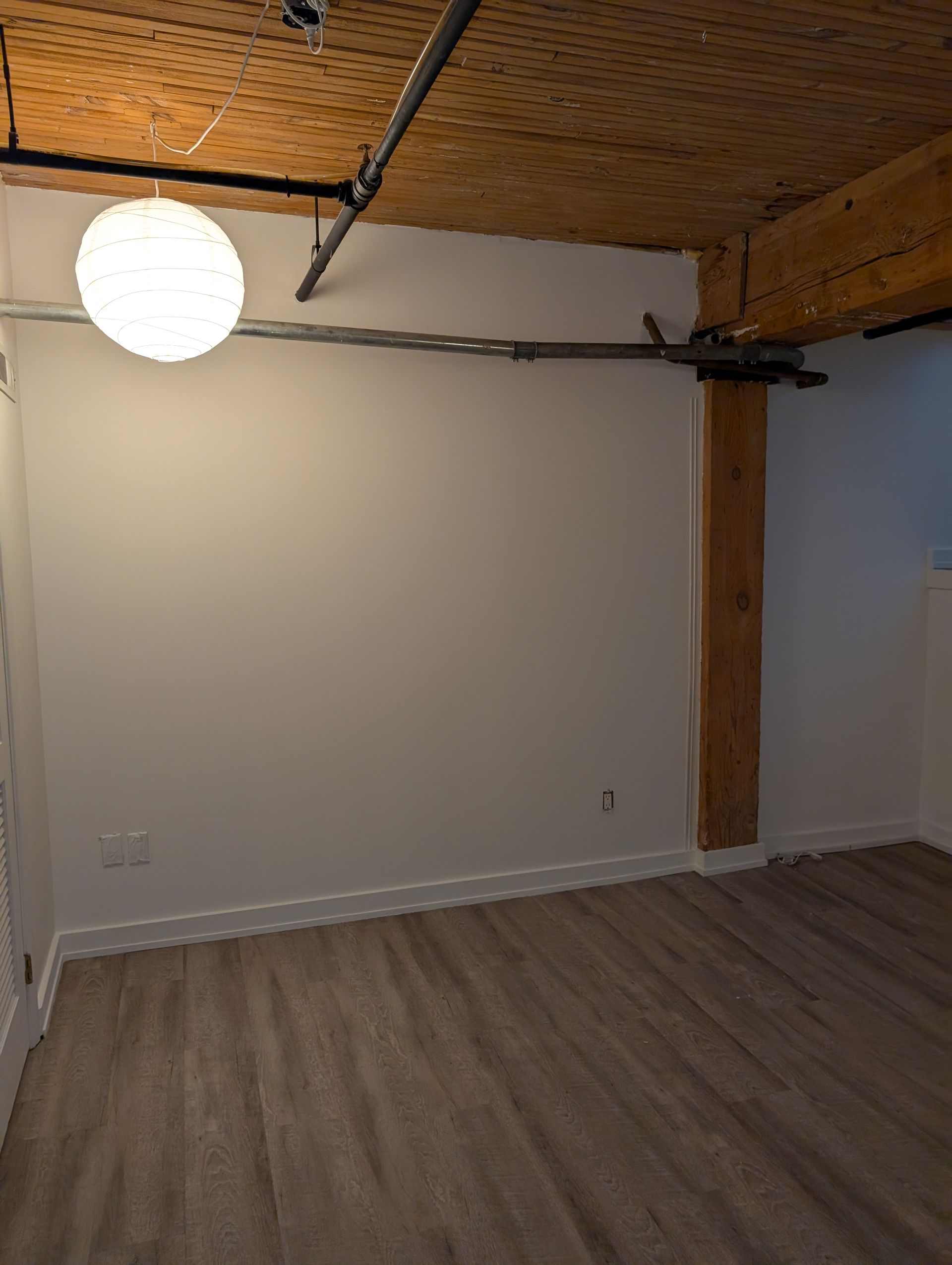 Empty room with white walls, wood floor, exposed wood ceiling, and a round hanging light fixture.