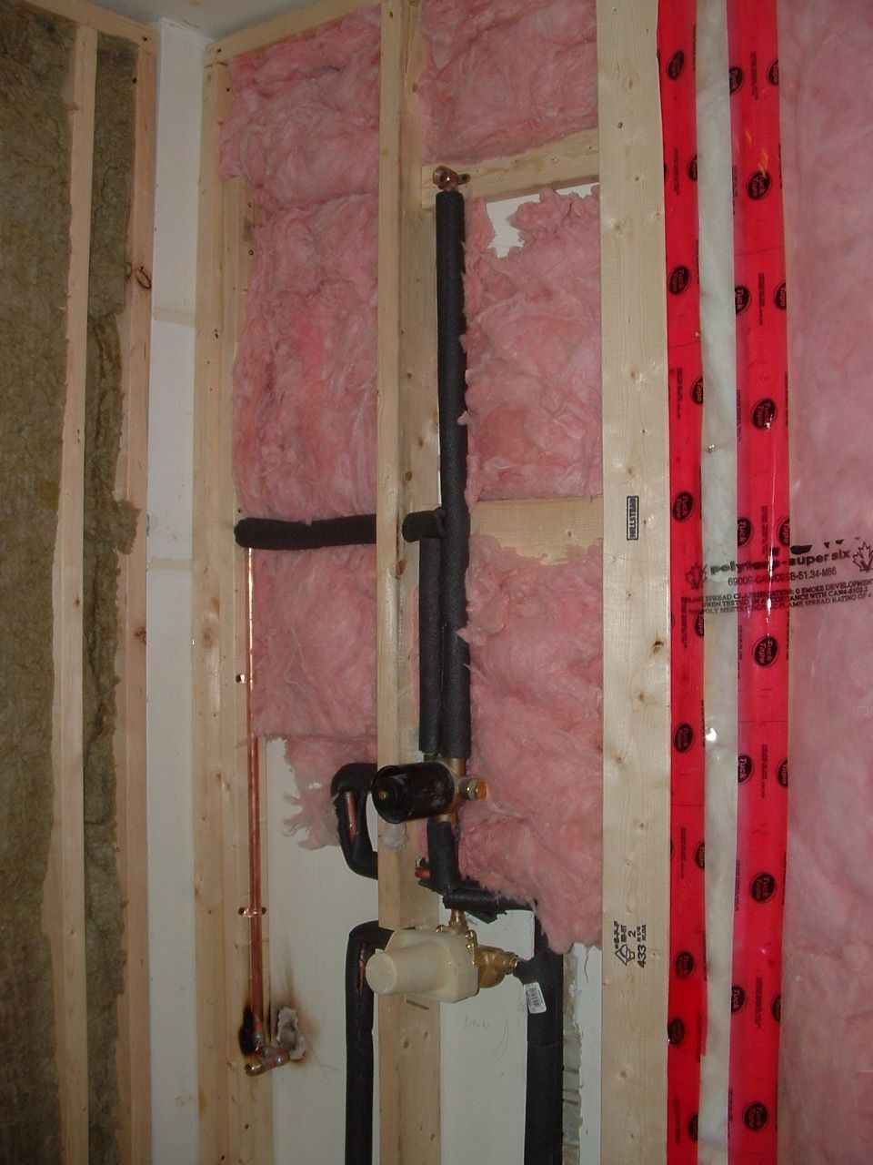 Exposed wall framing with pink insulation and plumbing pipes inside a room under construction