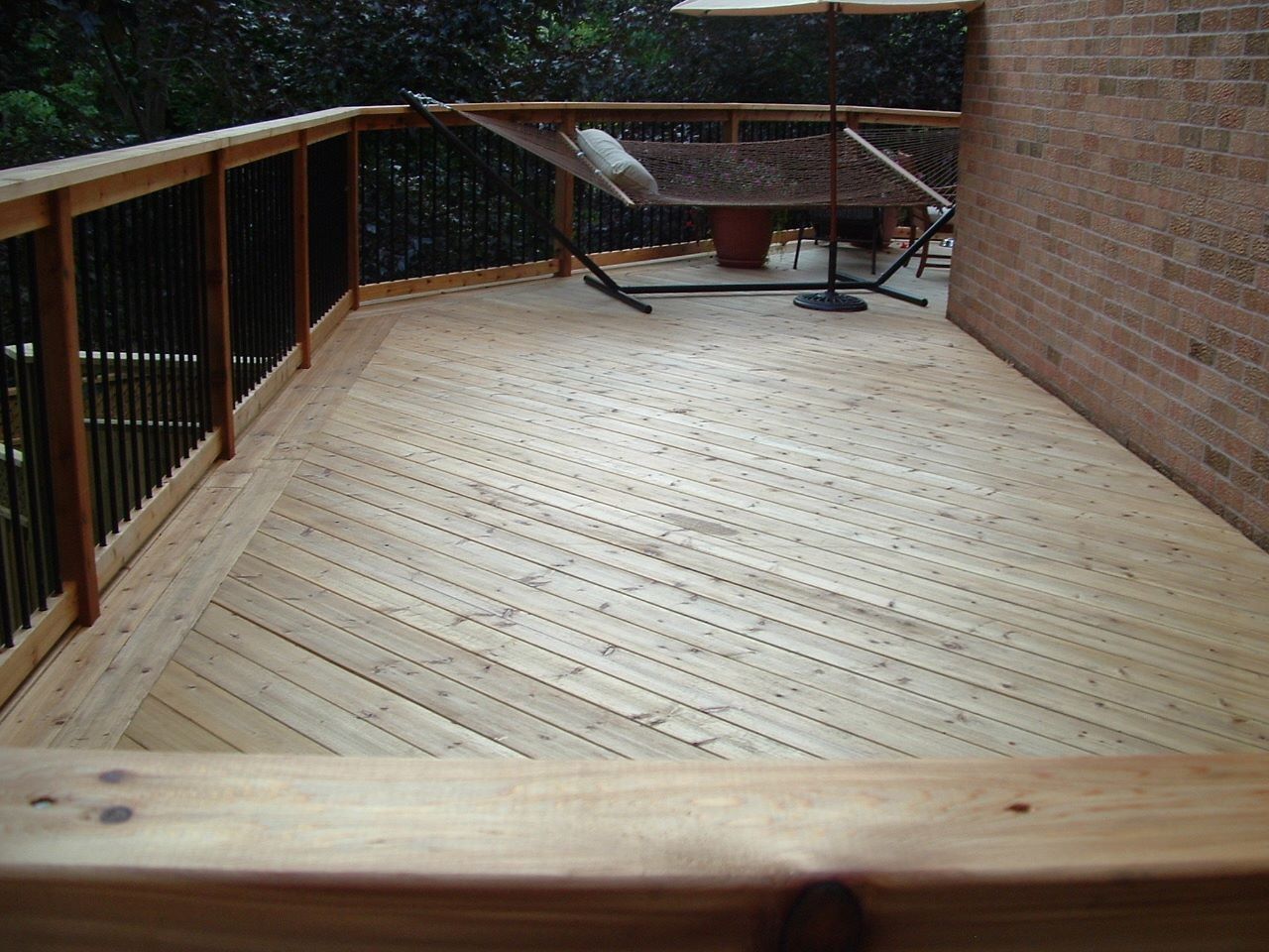 Wooden deck with railing beside a brick wall, viewed from one end toward a yard and patio furniture.