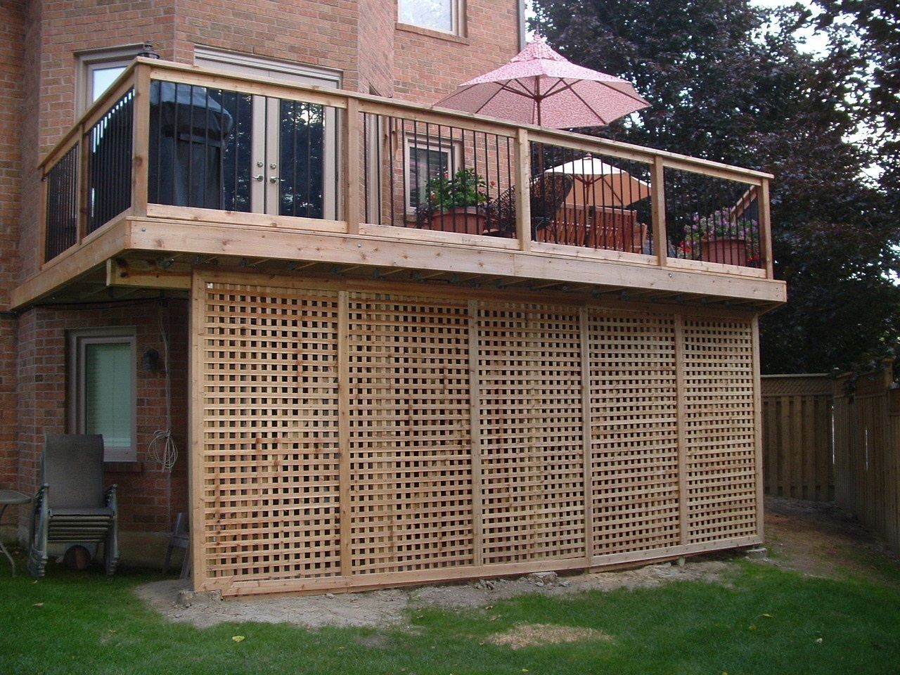 Backyard deck atop lattice-enclosed patio beside a brick house, with pink umbrella and potted plants.
