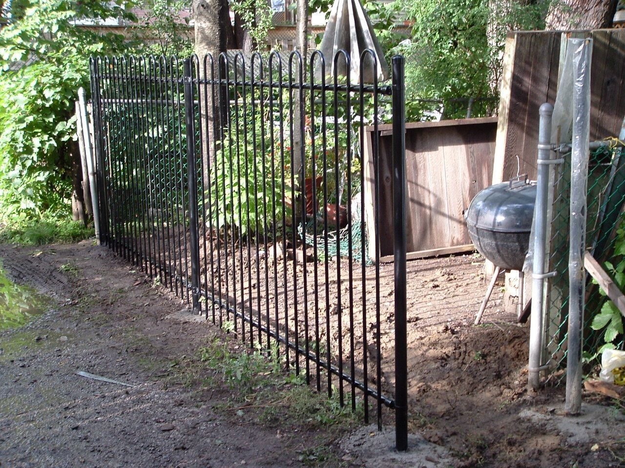 Black iron gate beside a dirt path and garden fence with a charcoal grill in the yard