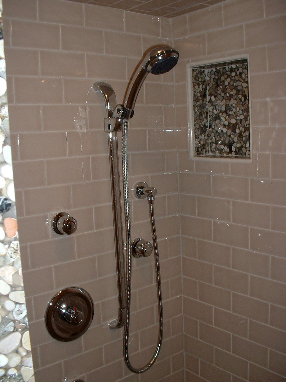 Chrome showerhead and controls on beige tiled bathroom wall with handheld sprayer and recessed niche.