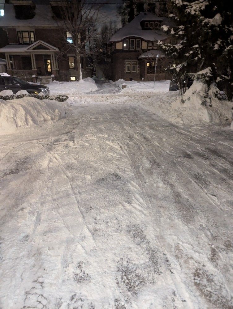 Snow-covered residential street at night with parked cars and houses in the background
