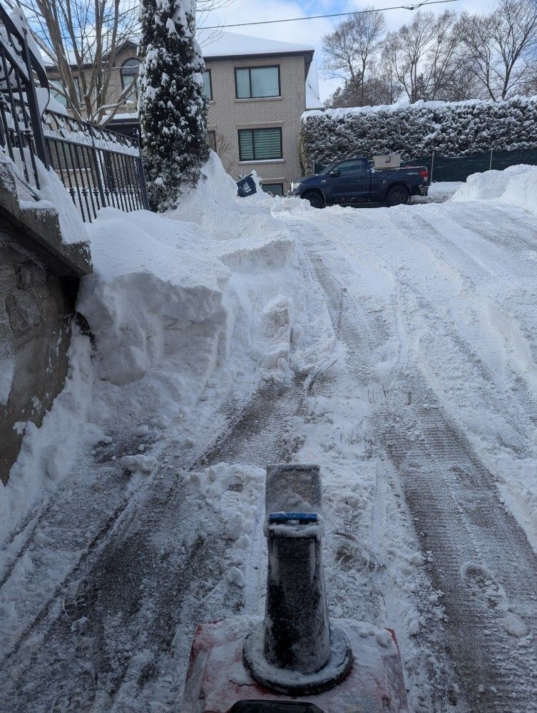 Snow-covered driveway with deep tire tracks and a black post in the foreground, apartment building in back