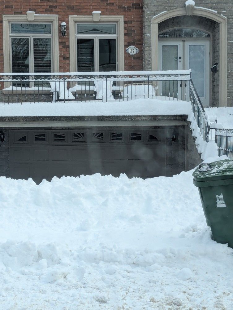 Snow-covered row house with a porch and stairs, and a green trash bin in front of a large snowbank.