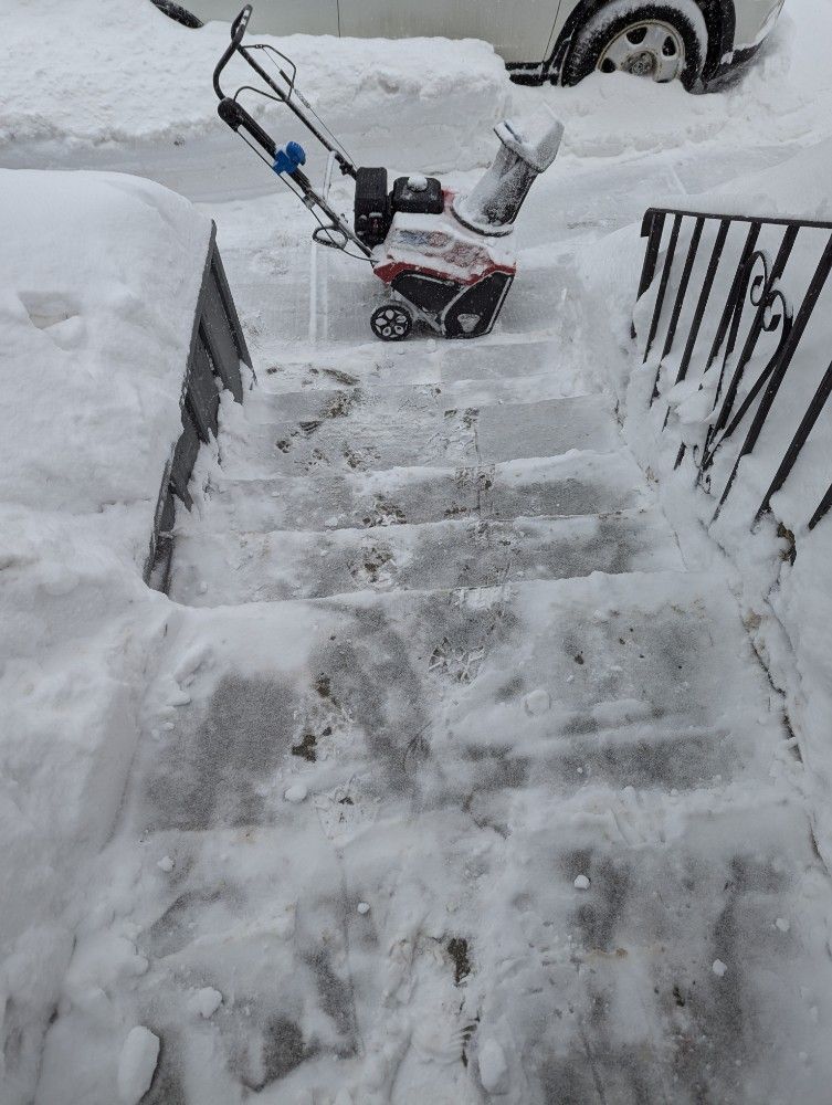 Snow-covered front steps leading to a driveway, with a snow blower at the top of the stairs.