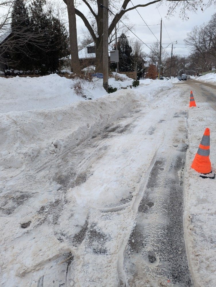 Snow-covered residential road with orange traffic cones and plowed tire tracks.
