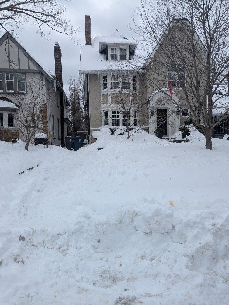 Snow-covered house with a large snowbank in front and bare trees on a winter street.