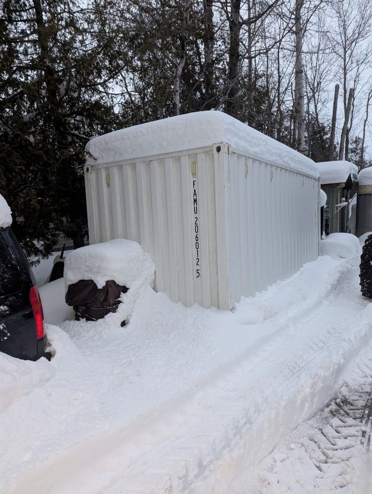 Snow-covered white shed beside a snowy road, with trees in the background.