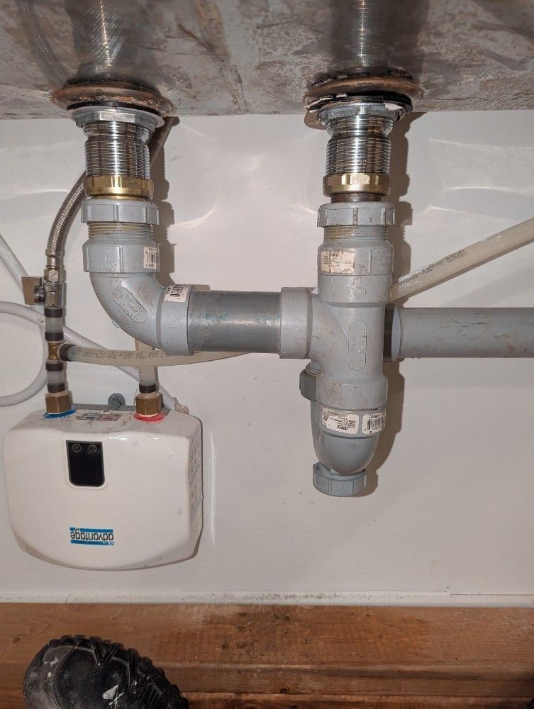 Under-sink plumbing with gray pipes and a small white water heater on a tiled wall