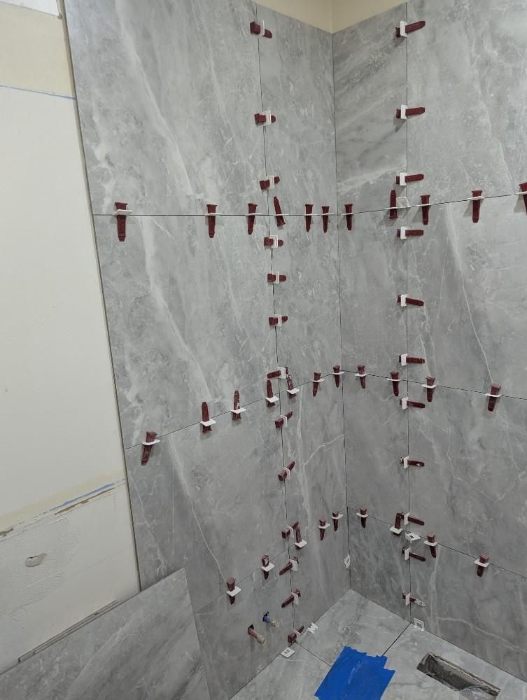Tile wall under construction with red leveling clips and spacers in a shower corner, blue tape on the floor