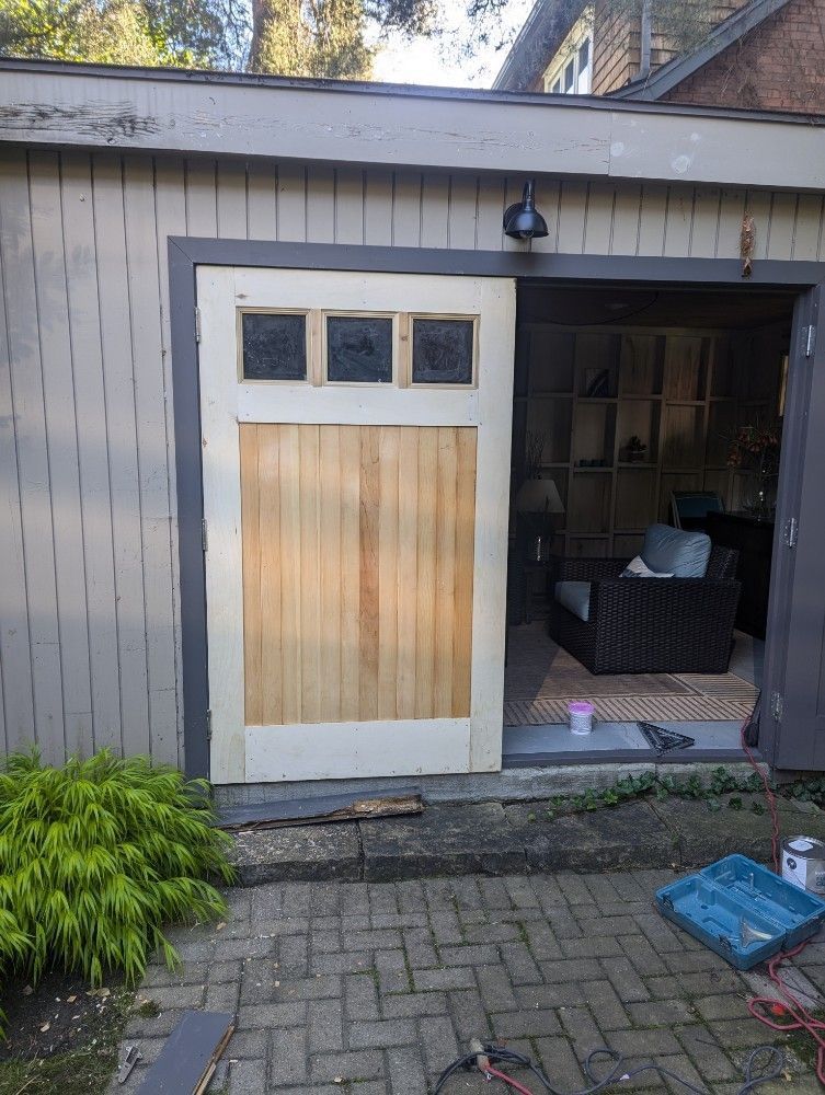 Open shed door with wicker chair inside beside a gray garage and brick patio.