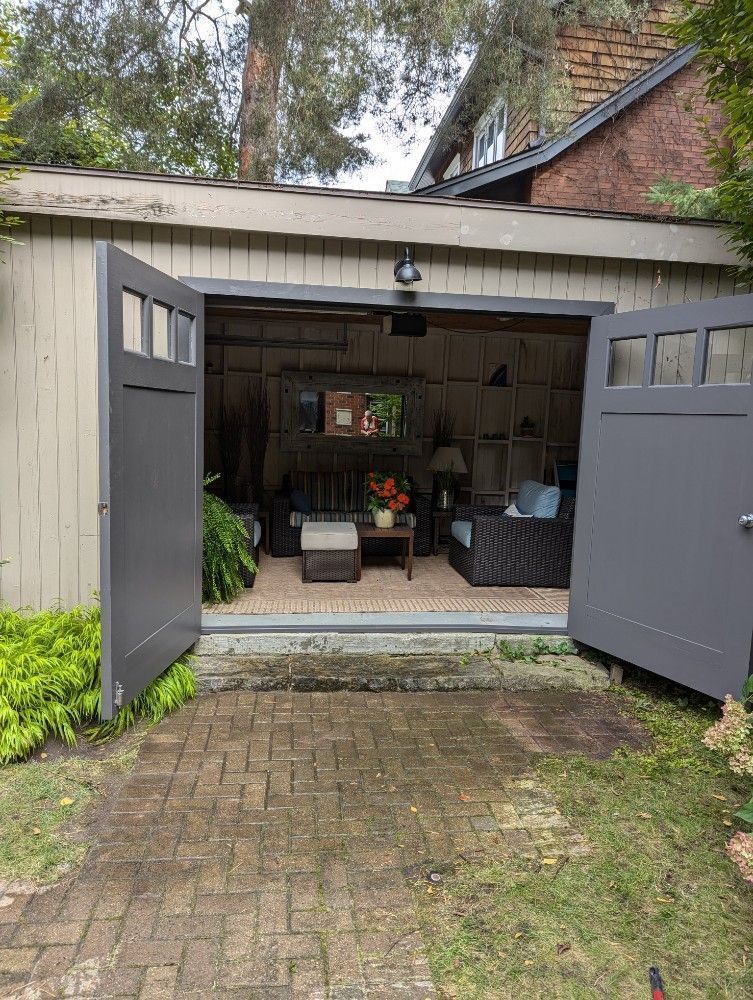 Open garage with double gray doors, revealing a cluttered interior and brick house behind.