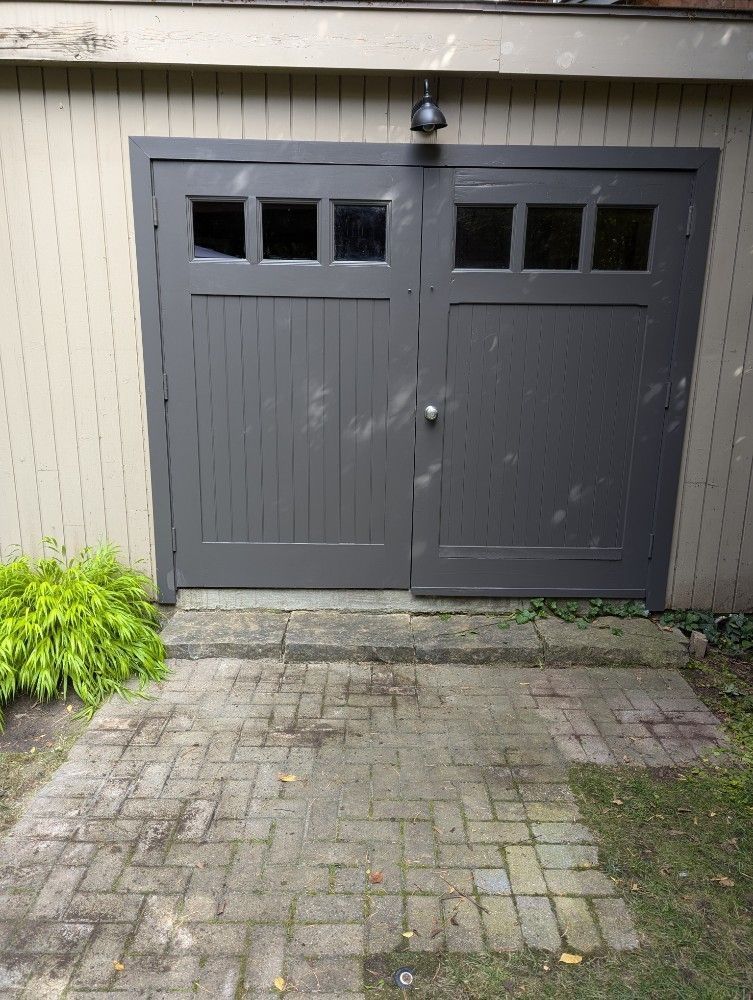 Dark gray double garage doors at the end of a brick driveway beside beige siding and plants