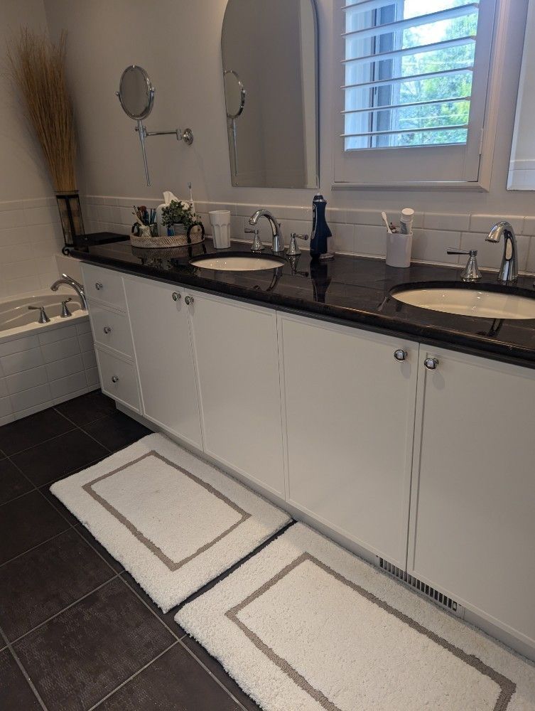 Bright white bathroom vanity with double sinks, black countertop, mirrors, and two white bath mats on dark tile floor