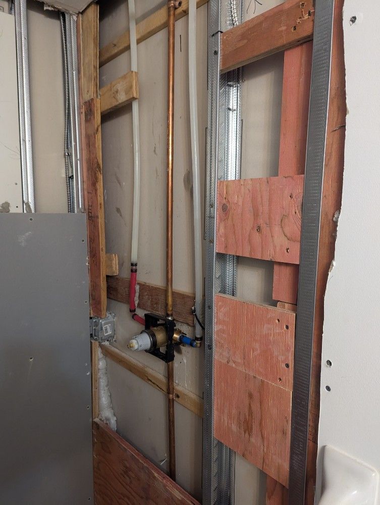 Partially renovated wall with exposed studs, pipes, and electrical wiring beside a gray panel.