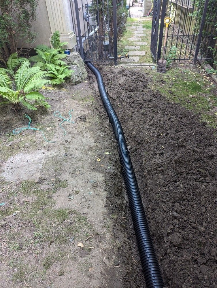 Garden trench with black corrugated pipe laid between a path and a soil bed near a gate.