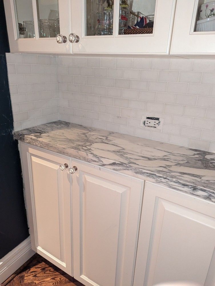 White kitchen cabinet with marble-look countertop and white tile backsplash under glass-front upper cabinets