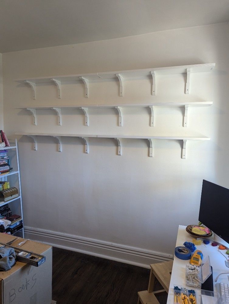 Empty white wall with three rows of white shelves above a baseboard heater and small desk area.