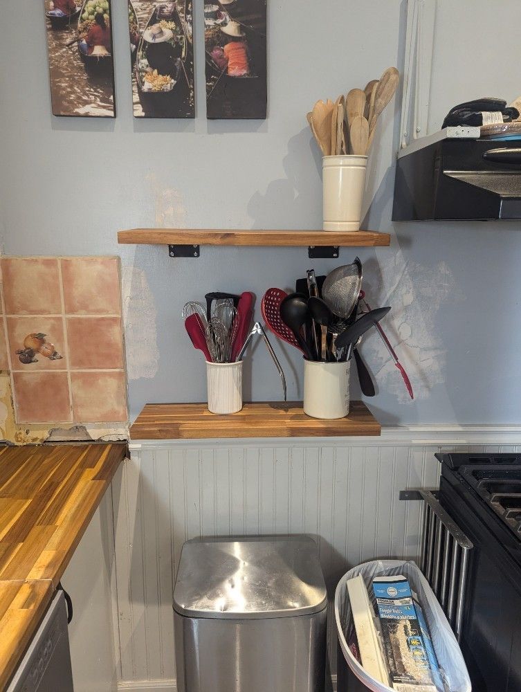 Kitchen counter with wooden shelf, white mugs of utensils, and metal storage rack above a sink area