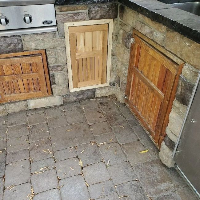 Stone patio corner with three small wooden cabinet doors beside a stainless steel grill and fridge