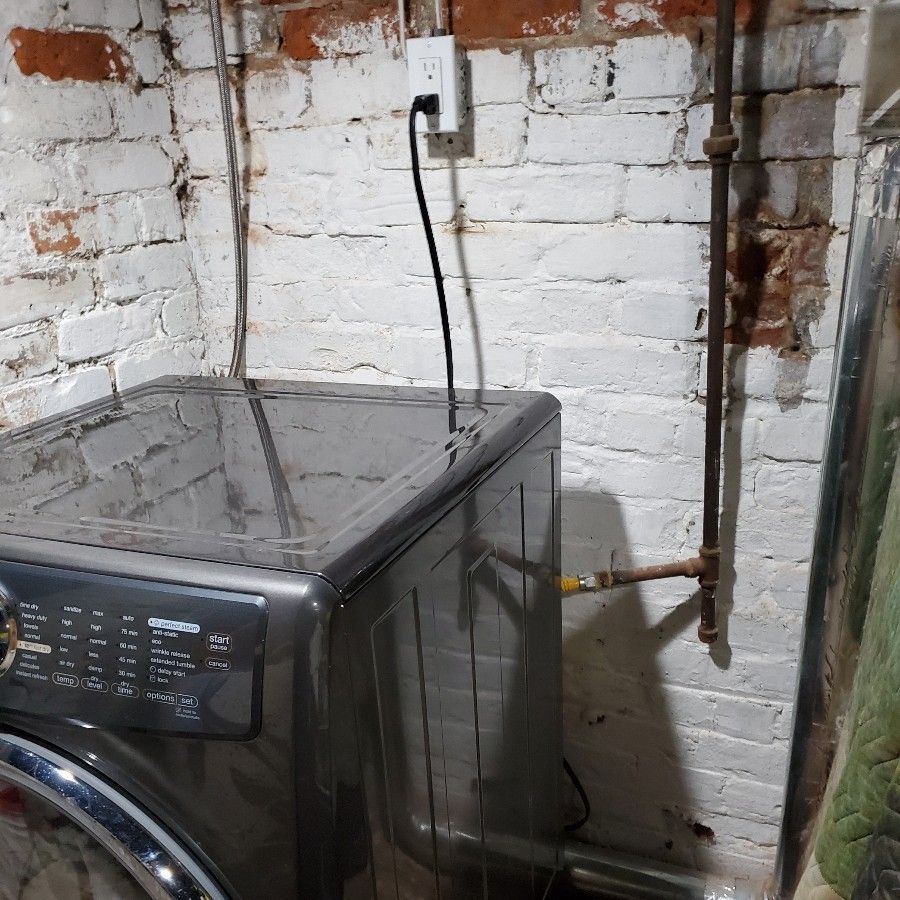 Front-loading washer beside a white brick wall with exposed plumbing and a wall outlet